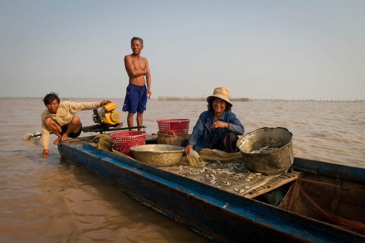 Three Cambodian fisherpeople on a long, narrow boat with fishing lines and baskets. They are on the rough, brackish water of Tonle Sap Lake.