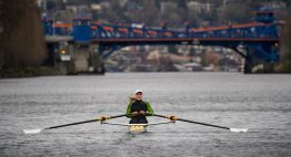 Female rower wearing a hat and wetsuit rows on Lake Washington with Fremont Bridge behind her.