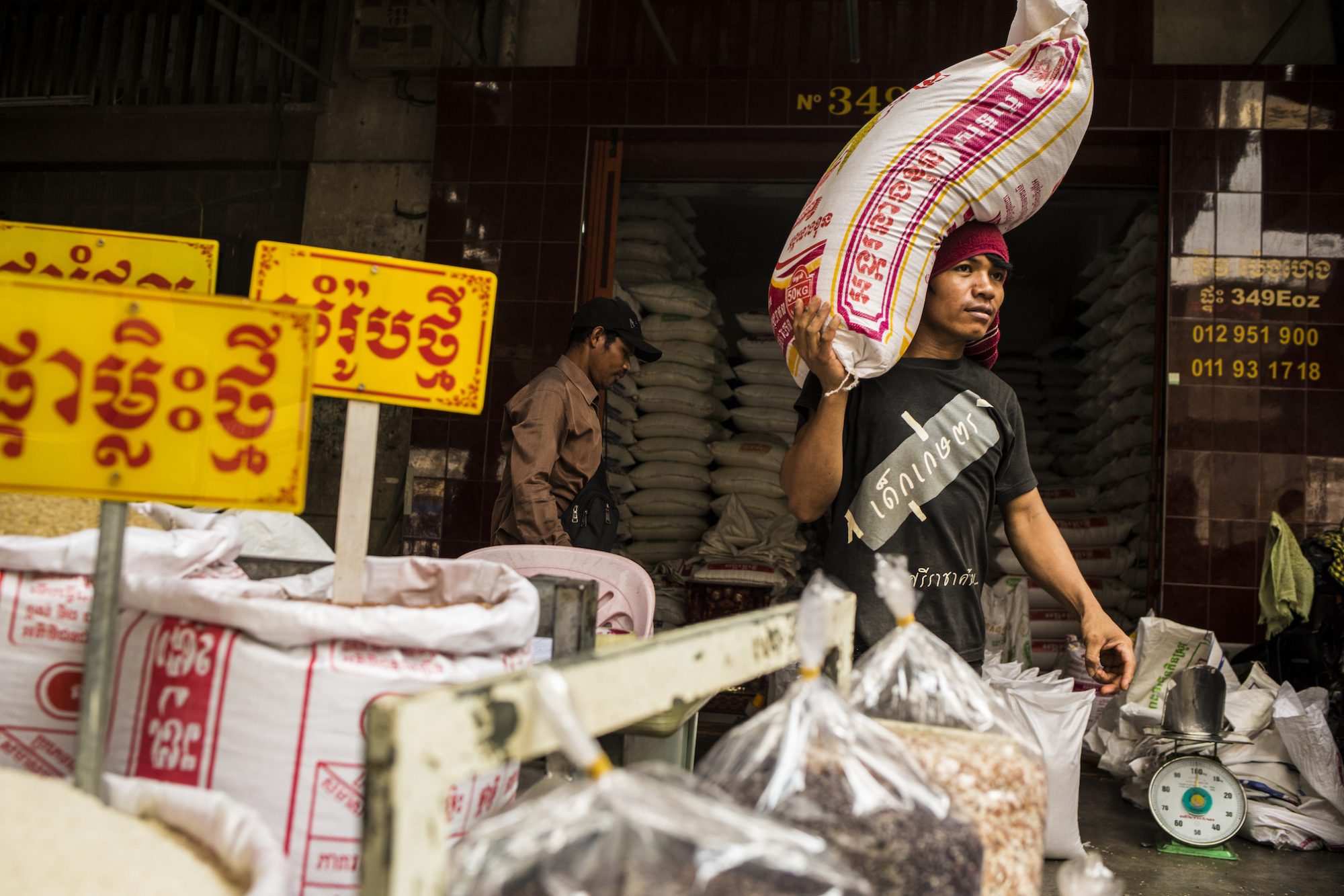 A man in a head scarf carries a huge bag of rice on his shoulder in a colorful rice market