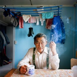 Older man sitting at kitchen table with a coffee mug and cigarette.