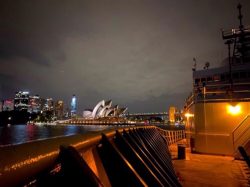 The deck of a ship with the Sydney Opera House lit at night.