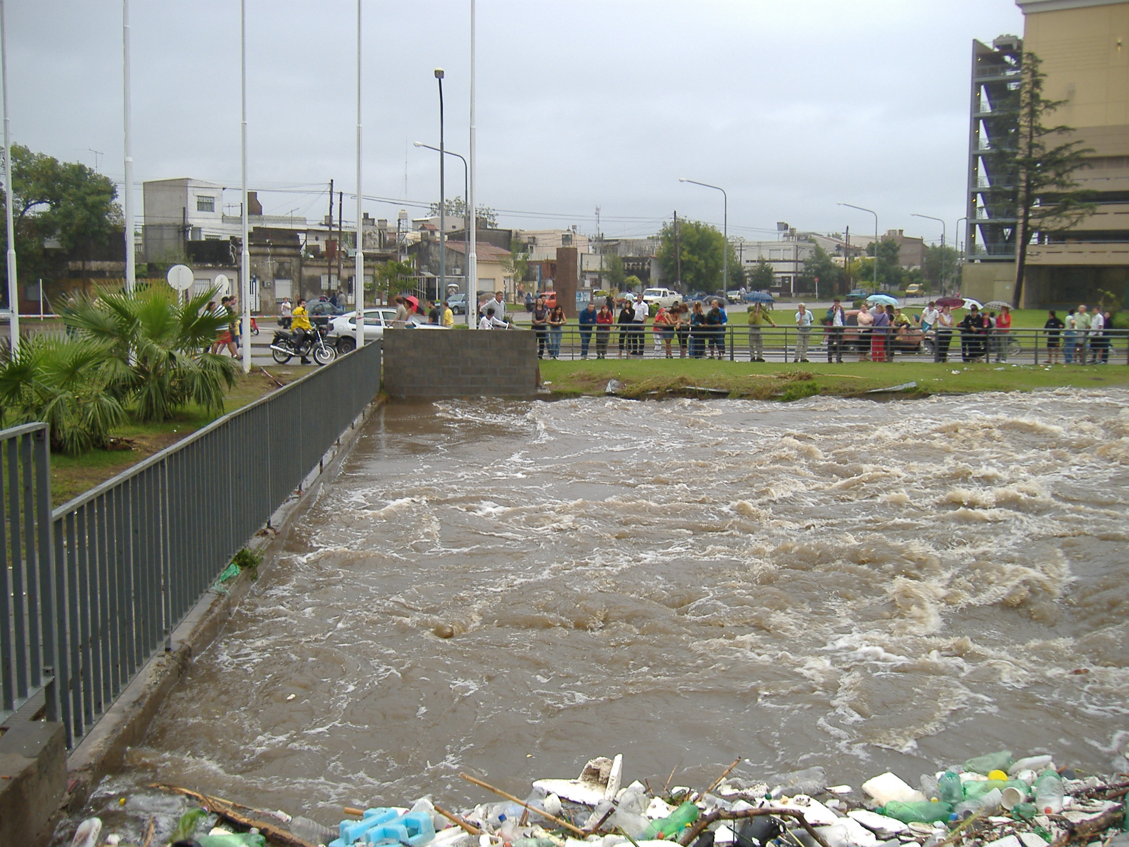 people gather on the edge of a flooding canal to look at the turbulent water
